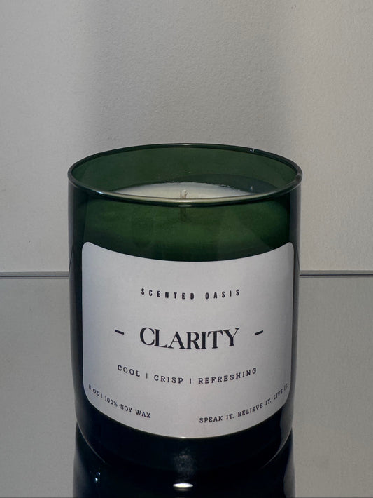 A dark green glass jar candle with a white label reading 'CLARITY' and 'SCENTED BAKS', photographed against a plain light gray wall and reflective surface.