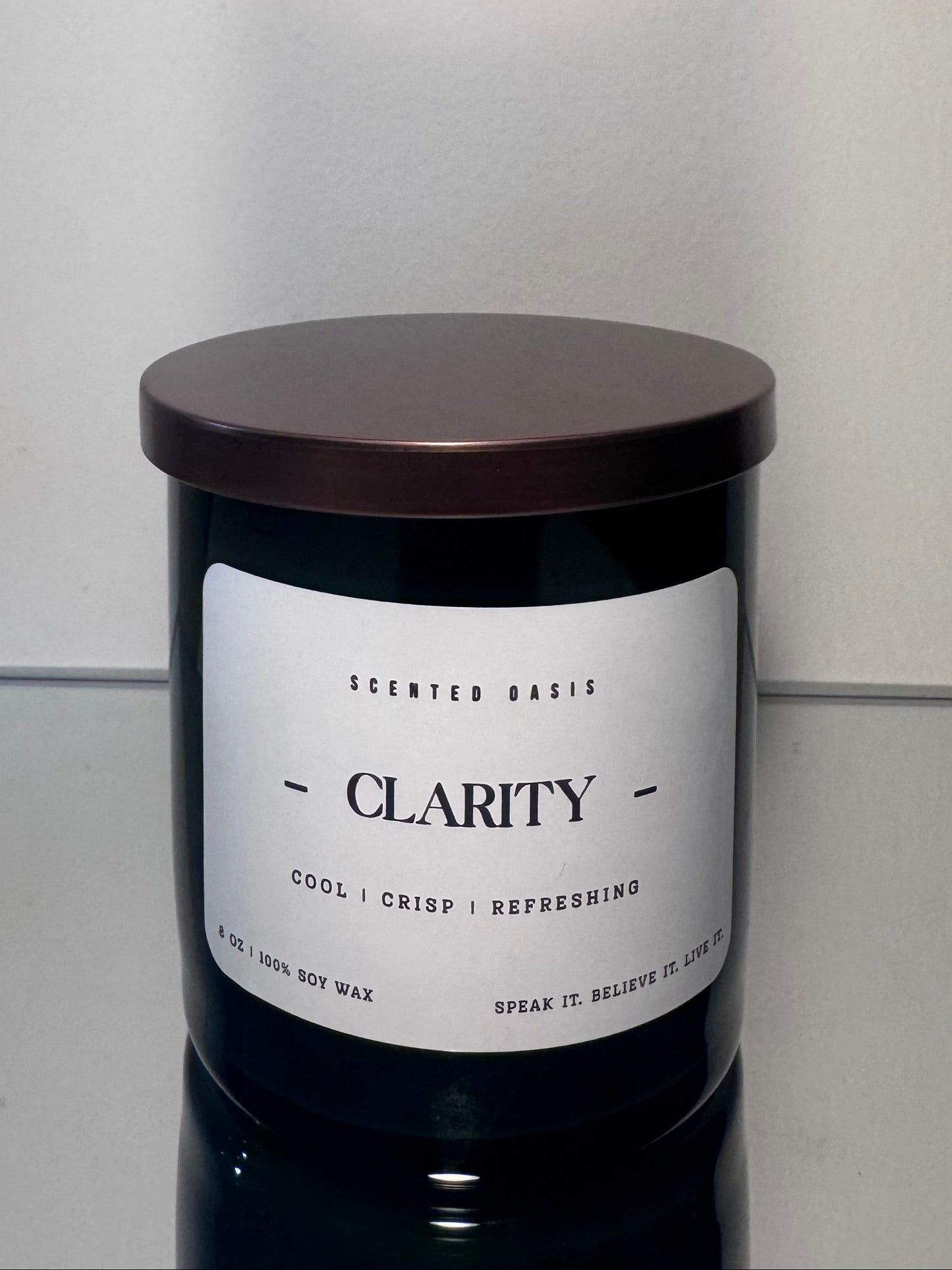 A dark glass jar candle with a white label reading 'CLARITY' and a dark brown lid, photographed against a plain light gray wall and surface.