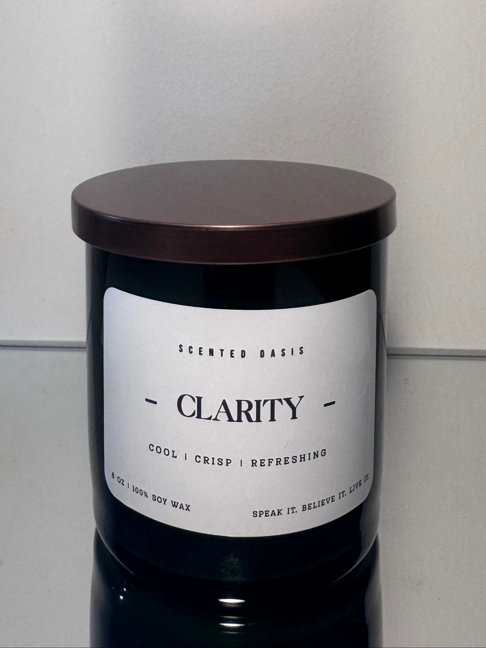 A dark glass jar candle with a white label reading 'CLARITY' and a dark brown lid, photographed against a plain light gray wall and surface.