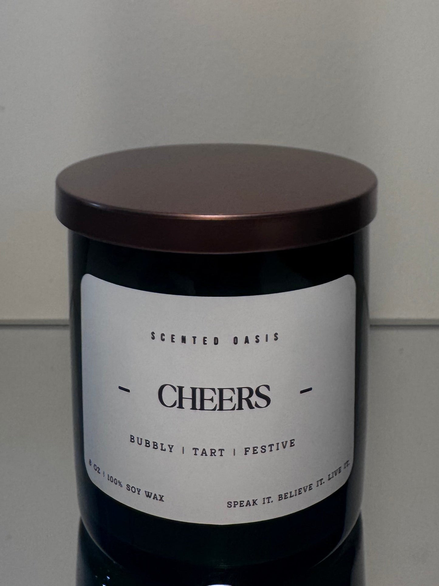 A dark glass jar candle with a metallic bronze lid and a white label reading 'CHEERS' and 'SCENTED BALSAM'.