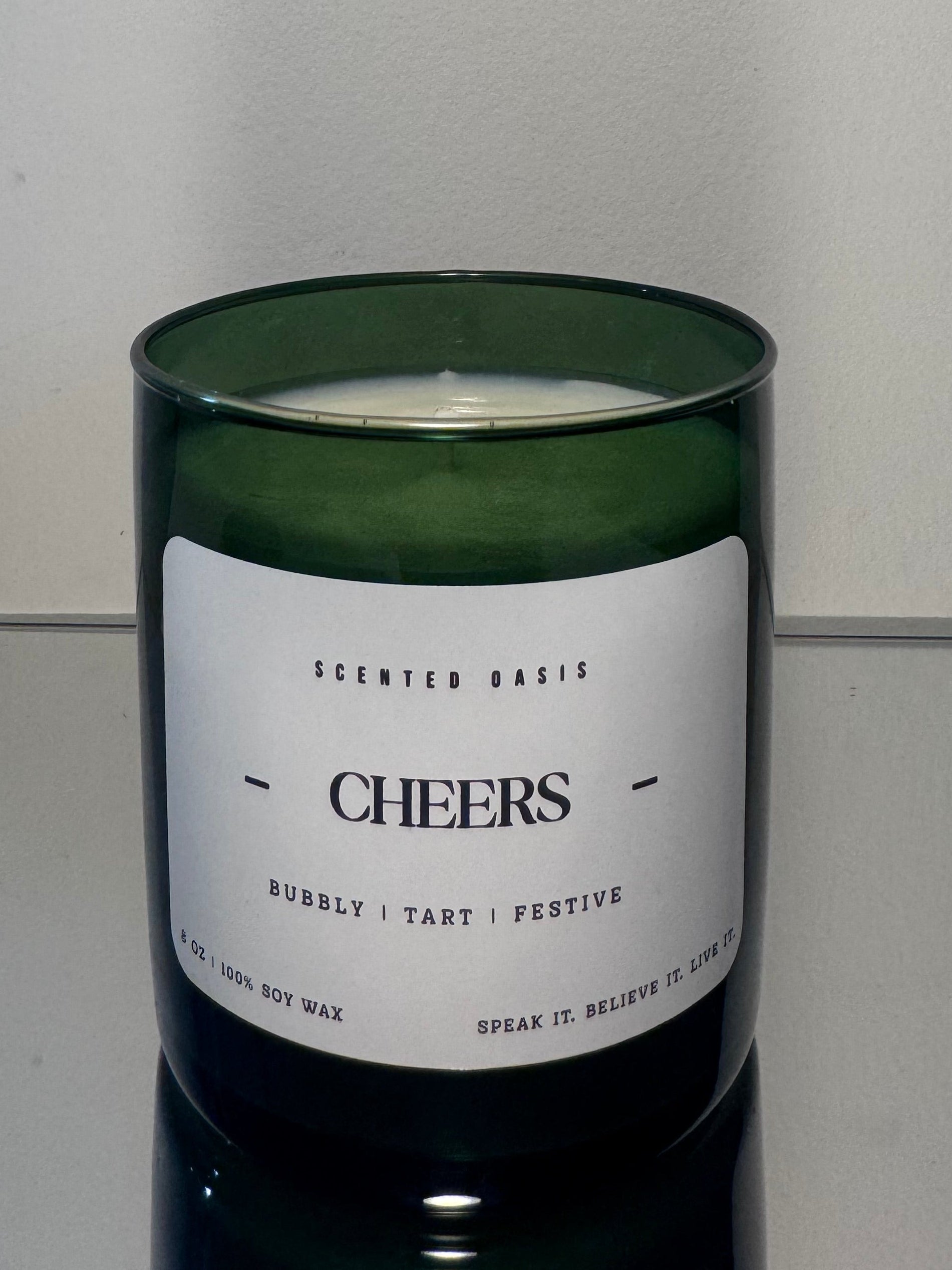 A green glass jar candle with a white label reading 'CHEERS' and smaller text 'SCENTED BASIS', photographed against a plain light background.