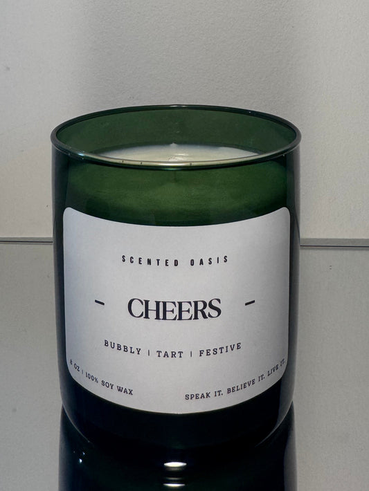 A green glass jar candle with a white label reading 'CHEERS' and smaller text 'SCENTED BASIS', photographed against a plain light background.