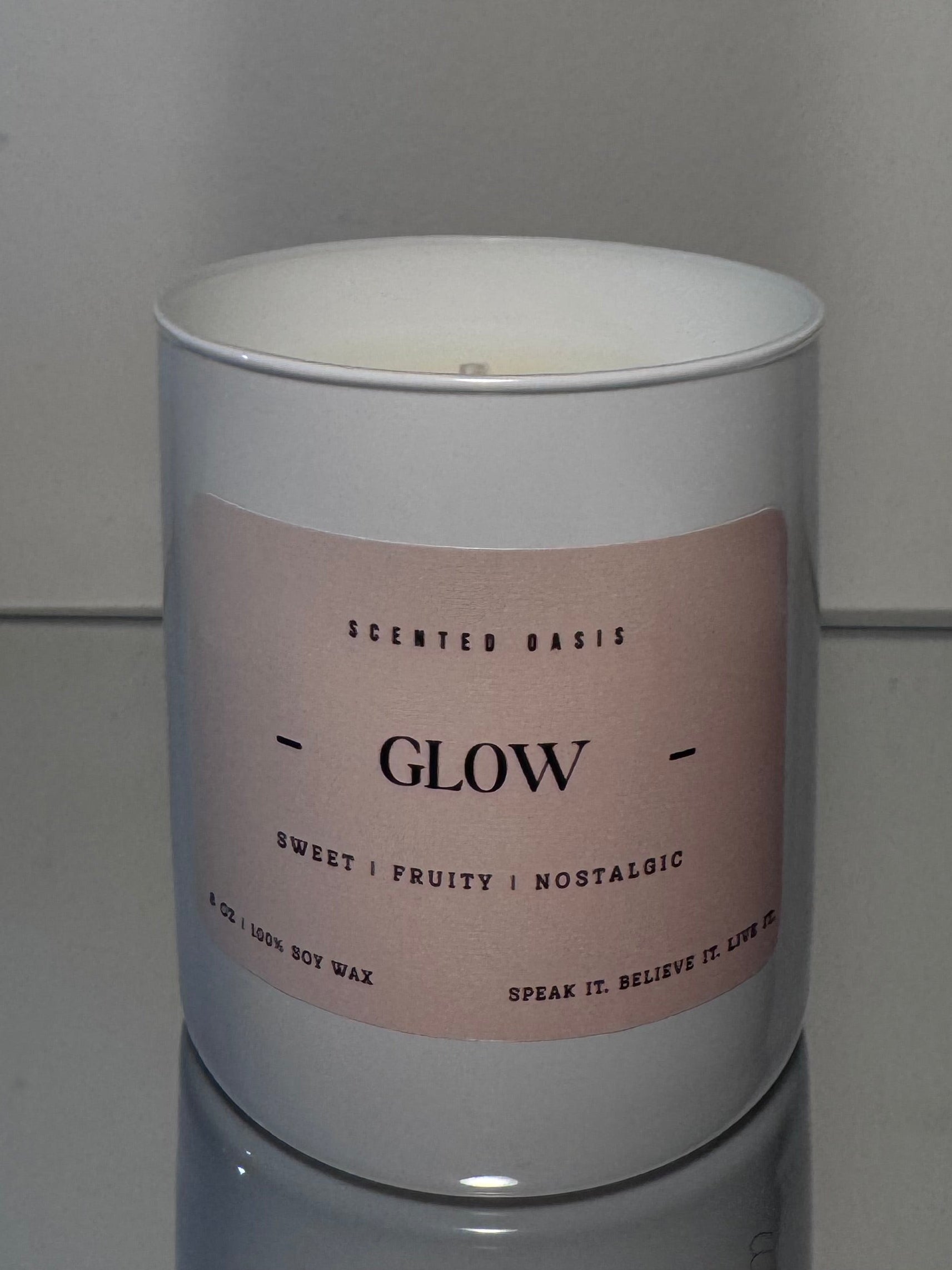 A white scented candle jar with a pale pink label reading 'GLOW' and smaller scent text, photographed against a plain light gray wall and reflective surface.