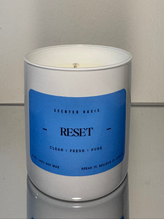 A white soy wax candle in a cylindrical white container with a blue label reading 'RESET' and smaller scent text, photographed against a plain light gray wall and surface.