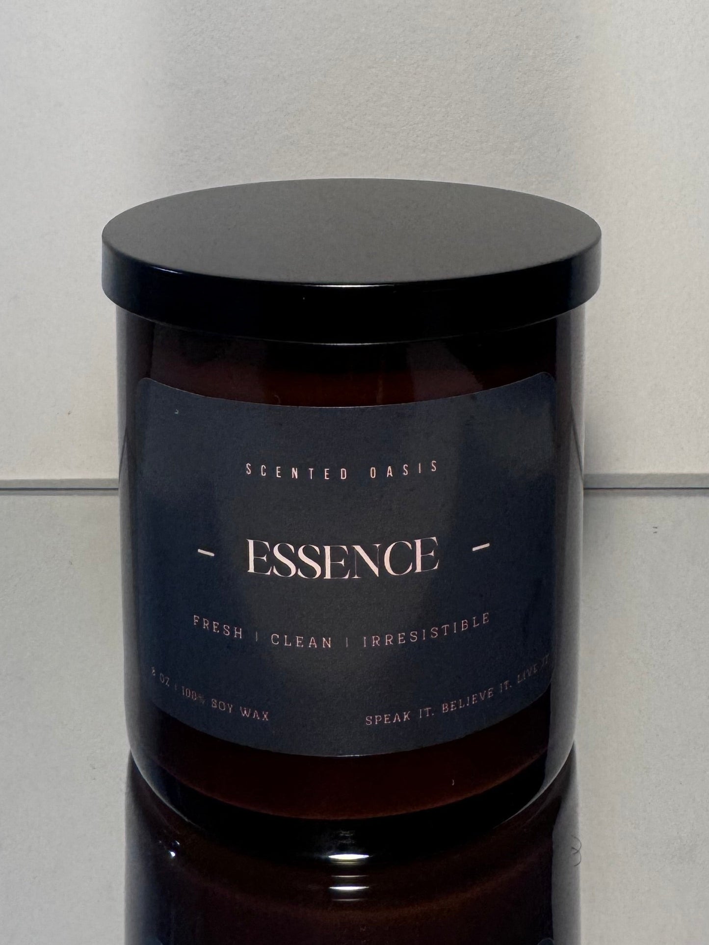 A dark amber glass jar candle with a black lid and a black label reading 'ESSENCE' and 'Scented Barks', set against a plain light gray background.
