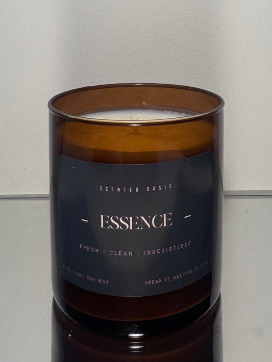 An amber glass jar candle with a dark navy label reading 'ESSENCE' and 'Scented Barks', set on a reflective surface against a plain light gray background.